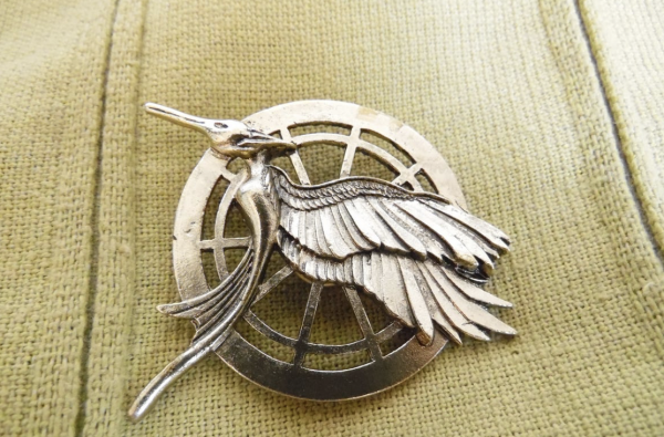 Pictured is a  mocking jay pin which is what Katniss Everdeen, the main character of the Original Hunger Games, wore. The Ballad of Songbirds and Snakes was a prequel to the Hunger Games.