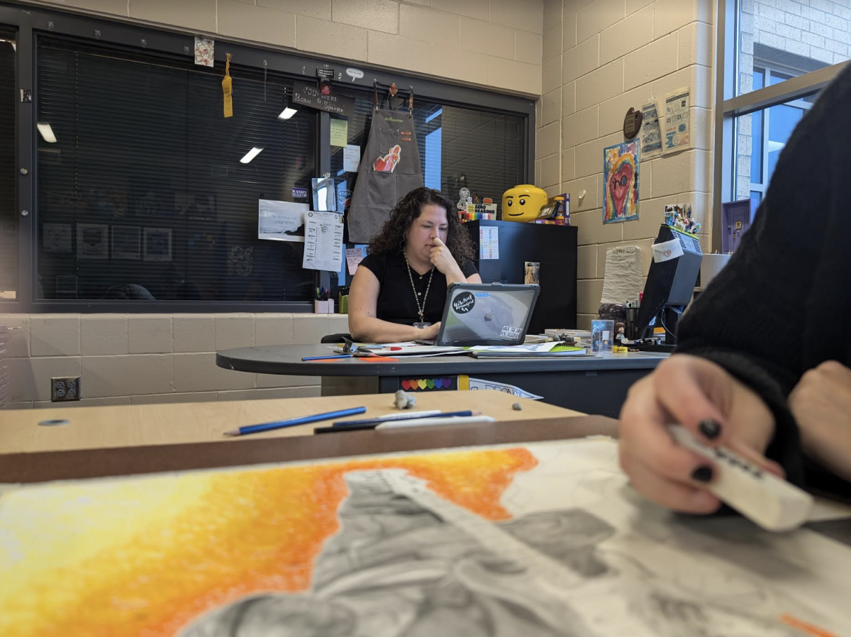 Abby Stallbaumer, art teacher, sits at her desk while her drawing class works. Students seek assistance with their projects. (Photo by A. Rushing). 