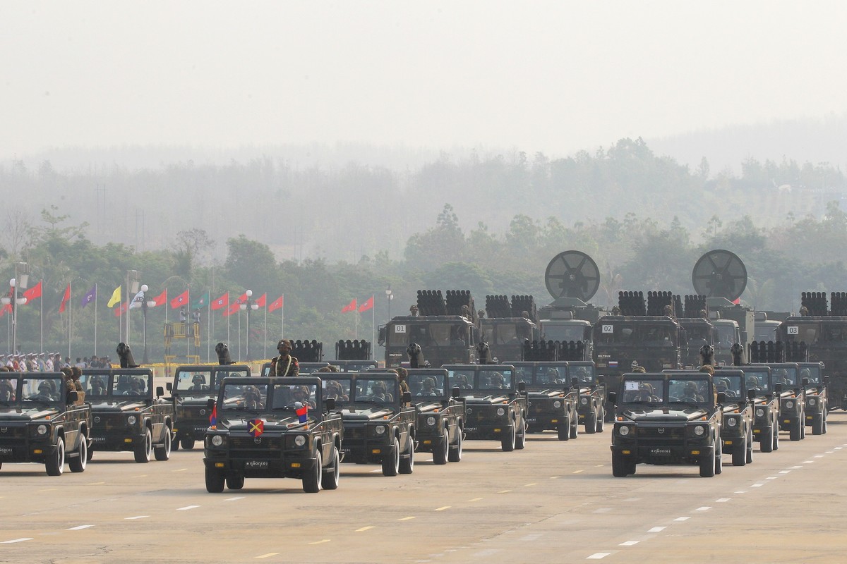 Myanmar military vehicles take part in a parade featuring soldiers and armored trucks during a national event, with rows of flags visible in the background. 