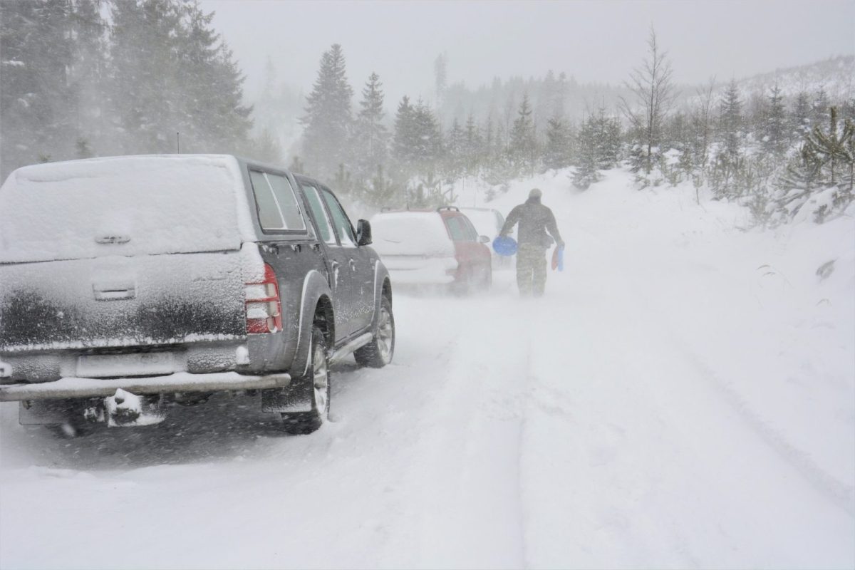 Cars sit along a snow-covered road as blowing snow and low visibility make travel dangerous. A person walks through the storm carrying supplies, showing the harsh conditions caused by severe winter weather.